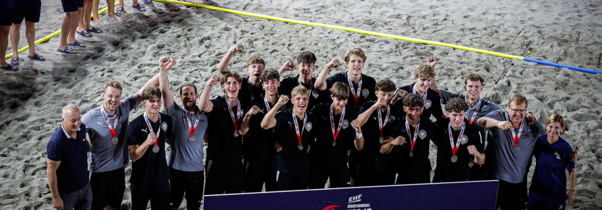 A group of athletes and staff celebrate on a sandy beach, wearing medals and posing with a sign that reads "2nd PLACE" at the EHE Beach Handball Euro 2026.