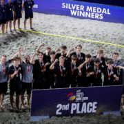 A group of athletes and staff celebrate on a sandy beach, wearing medals and posing with a sign that reads "2nd PLACE" at the EHE Beach Handball Euro 2026.
