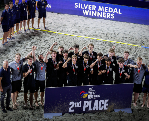 A group of athletes and staff celebrate on a sandy beach, wearing medals and posing with a sign that reads "2nd PLACE" at the EHE Beach Handball Euro 2026.