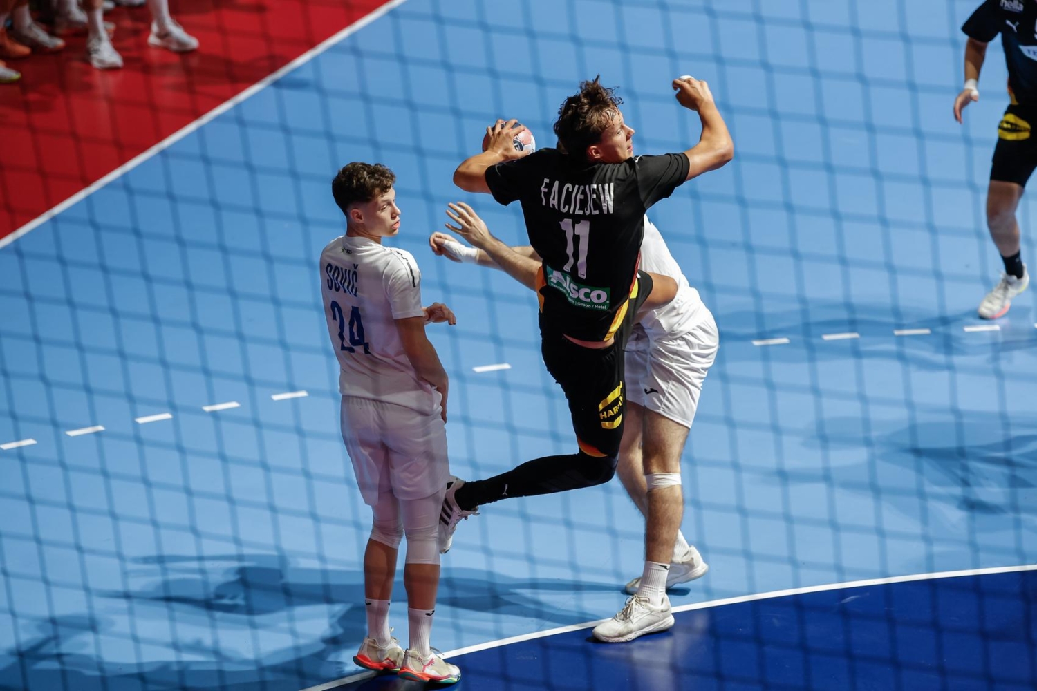 A player in a black jersey with the number 11 leaps to strike a ball during a handball match, while an opponent in a white jersey watches closely.