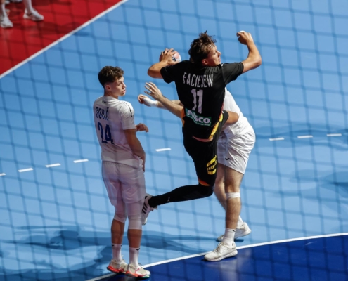 A player in a black jersey with the number 11 leaps to strike a ball during a handball match, while an opponent in a white jersey watches closely.