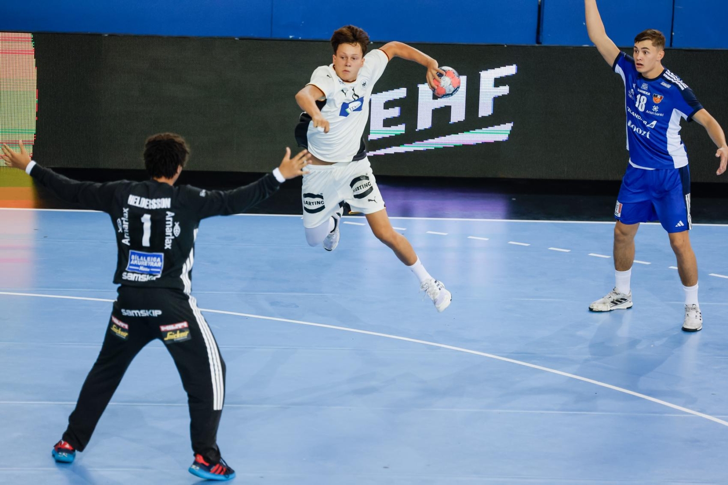 A player in a white uniform leaps to pass the ball while being defended by an opponent in black, with another player in blue observing from the side.